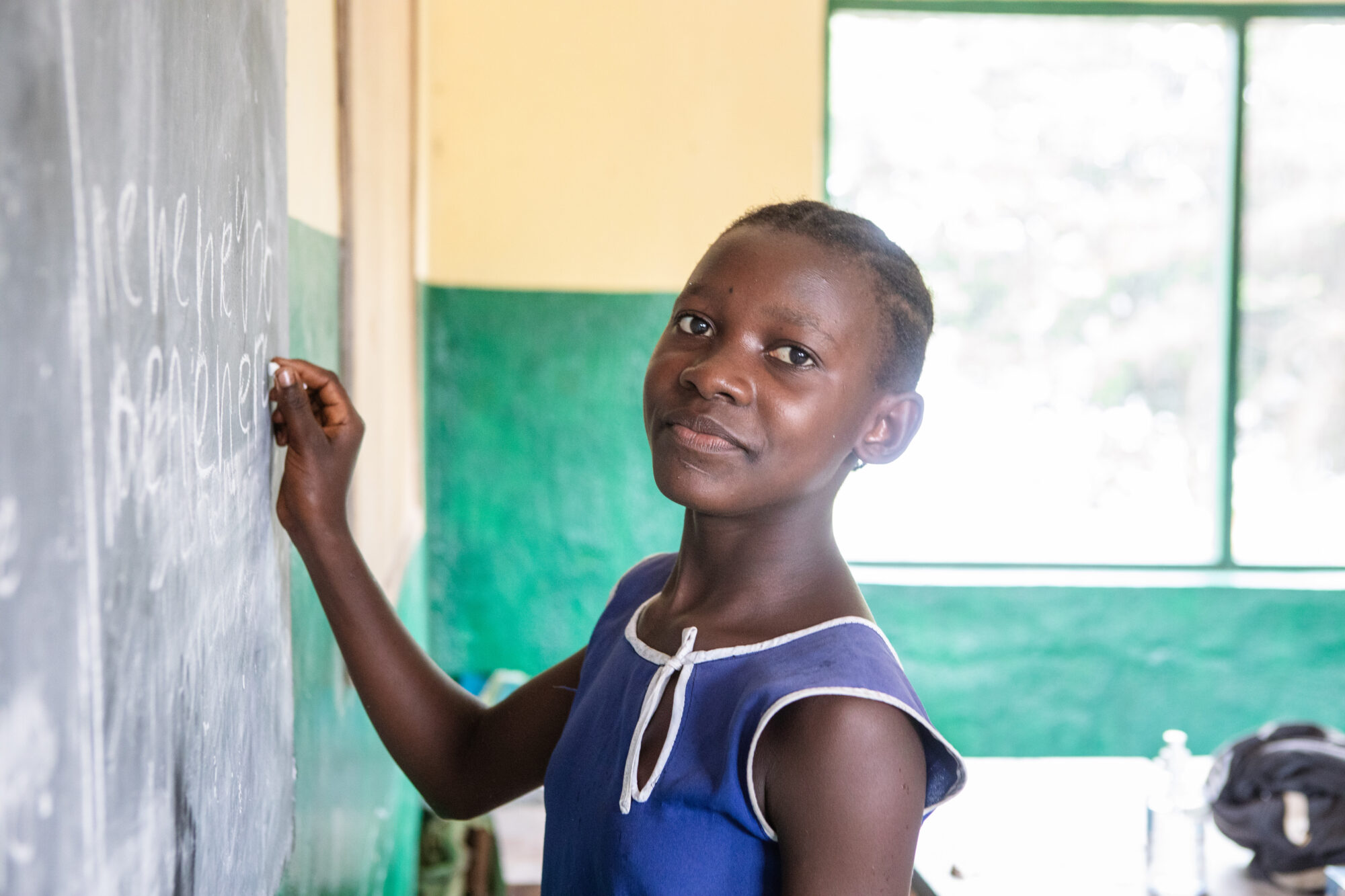 Tenneh age 13 at her school, girl smiling and writing on chalkboard