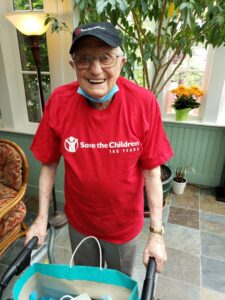 John Hillman, a WWII veteran, does an annual walkathon, with funds supporting children and families in crisis around the world. He is posing with his walker, wearing a hat and shirt from Save the Children. 