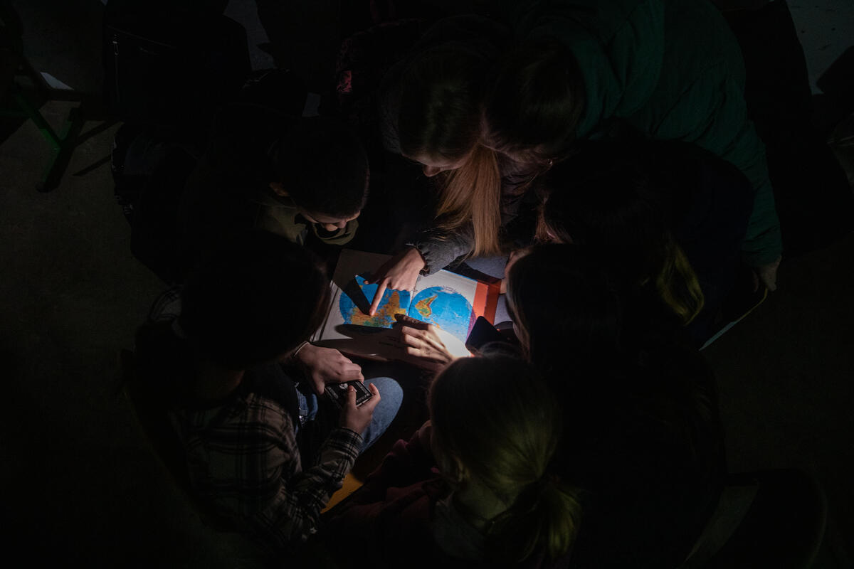 Children study geography in a school basement during air alert in Chernihiv region, Ukraine. With no electricity, water, or heating students must use cellphones to see their textbooks.