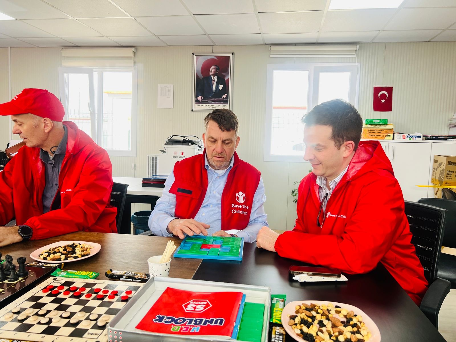 Danny (right) sits at a table with two other male Save the Children staff members inside a container classroom.