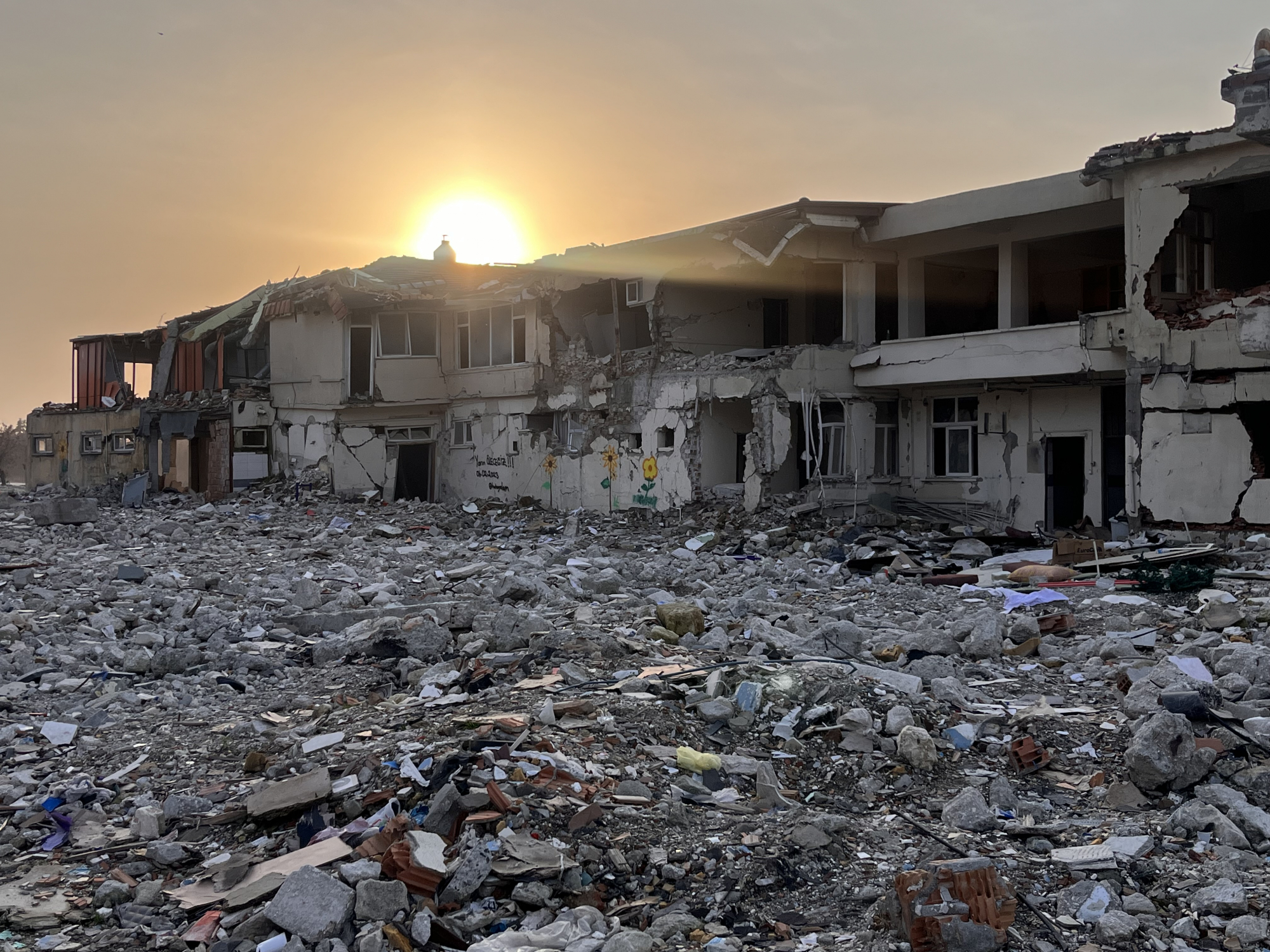 The sun rises over the remains of building destroyed by the earthquake. In the foreground is a pile of rubble from the damaged building.